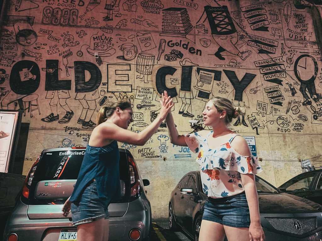 blonde and brunette girl high fiving in front of old city mural