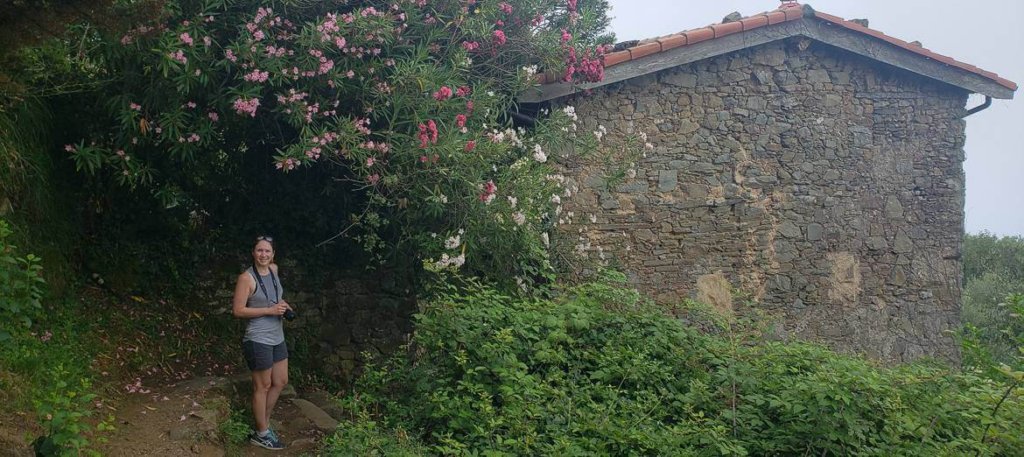 brunette girl in gray tanktop in front of stone building surrounding by flowering greenery