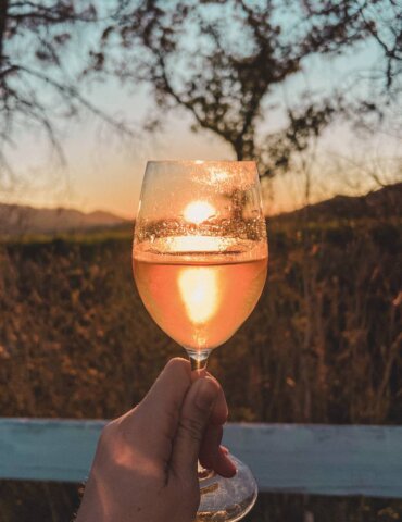 Hand holding wine glass in front of setting sun with vineyard in the background - napa valley itinerary