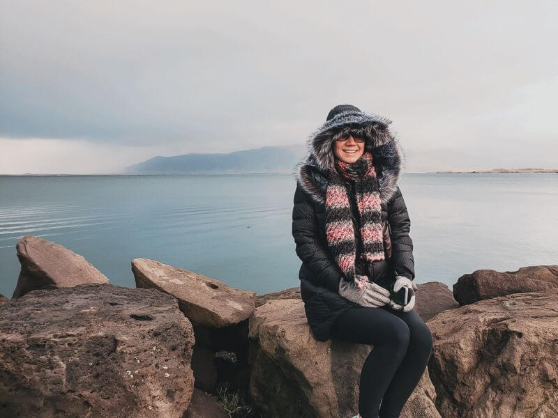 girl wearing parka and sitting in front of ocean in Reykjavik
