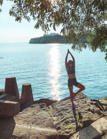 brunette girl in black yoga clothes doing tree pose near a lake