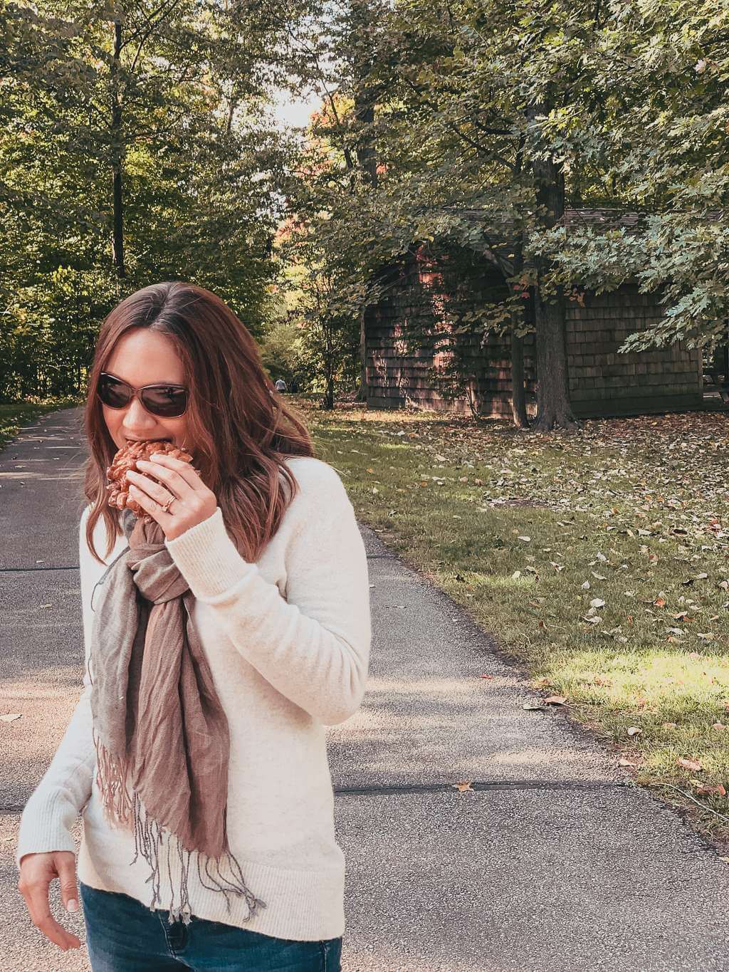 girl in white sweater, pink scarf, and sunglasses eating apple fritter along path in october
