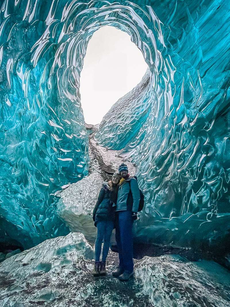 Couple standing near opening in ceiling of blue ice cave