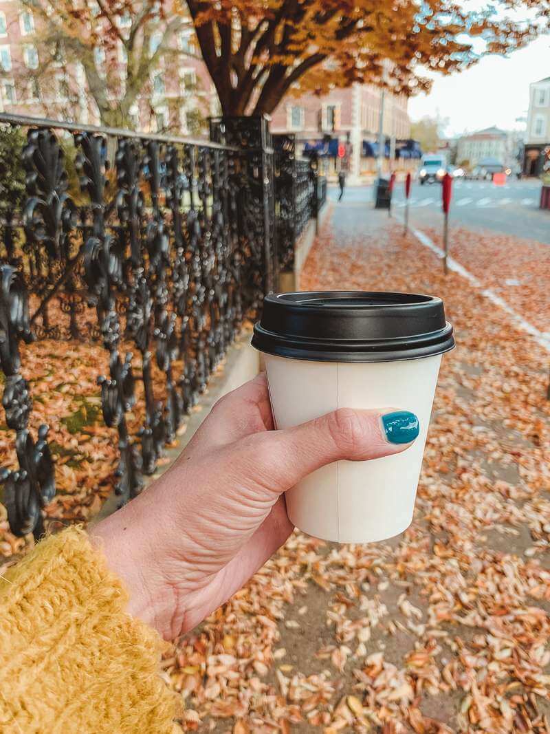 Girl in yellow sweater holding paper takeaway cup of coffee on crisp fall day with leaves everywhere - trip to Salem MA