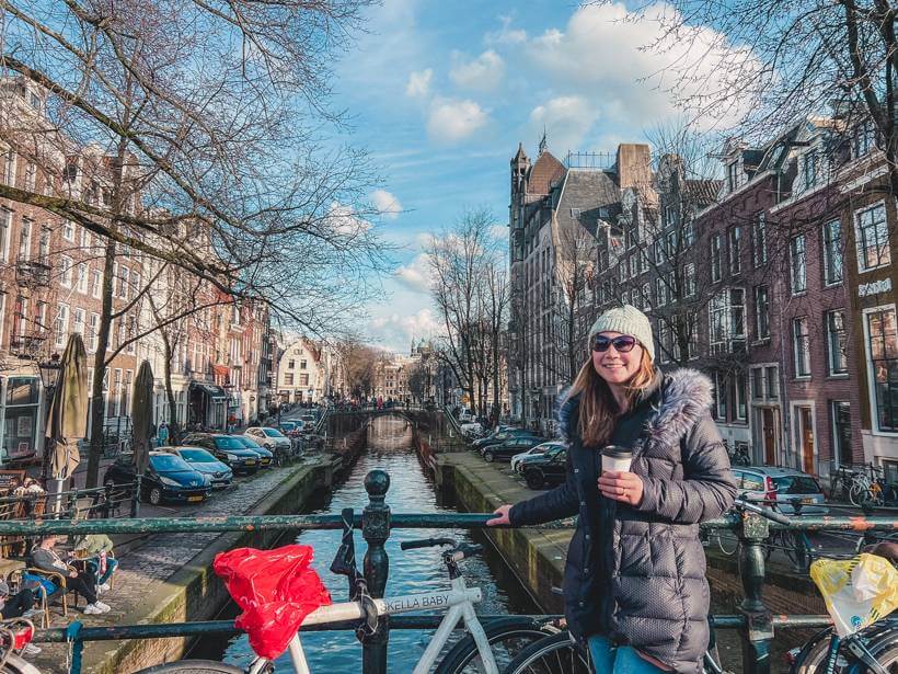 Girl wearing winter coat and knit hat holding coffee and posing in front of canal in Amsterdam