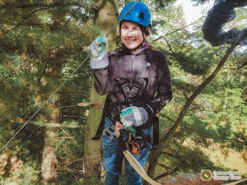 Girl in blue helmet, harness, fleece, blue athletic pants standing on platform in the trees