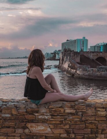 Girl sitting on wall outside Caribe Hilton during sunrise in Puerto Rico overlooking the Caribbean and a small fort.