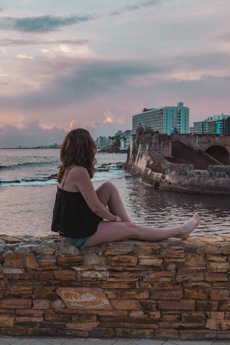 Girl sitting on wall outside Caribe Hilton during sunrise in Puerto Rico overlooking the Caribbean and a small fort.