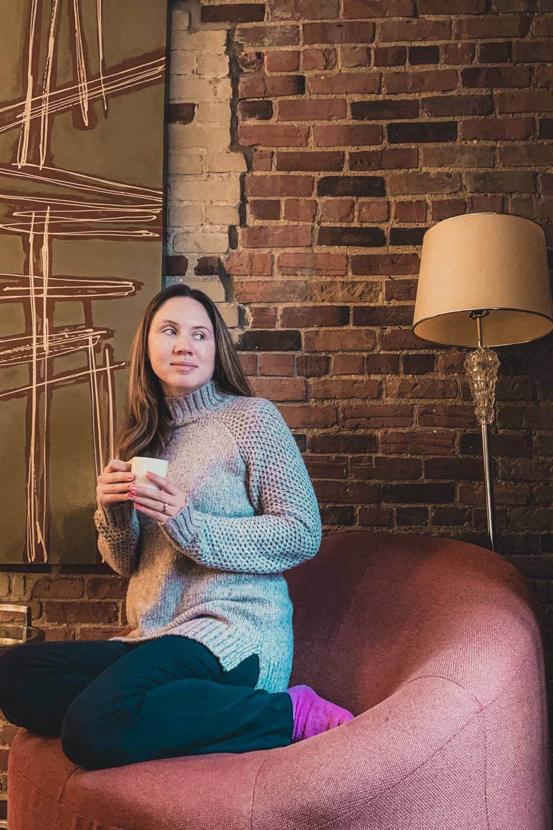 brunette girl in oversized sweater cozying up in hotel room with brick wall during solo travel in Montreal, sipping from a white mug of coffee