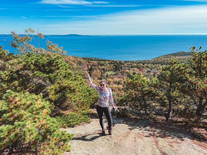 Girl standing with Atlantic Ocean and fall foliage in the background on the Beehive at Acadia National Park