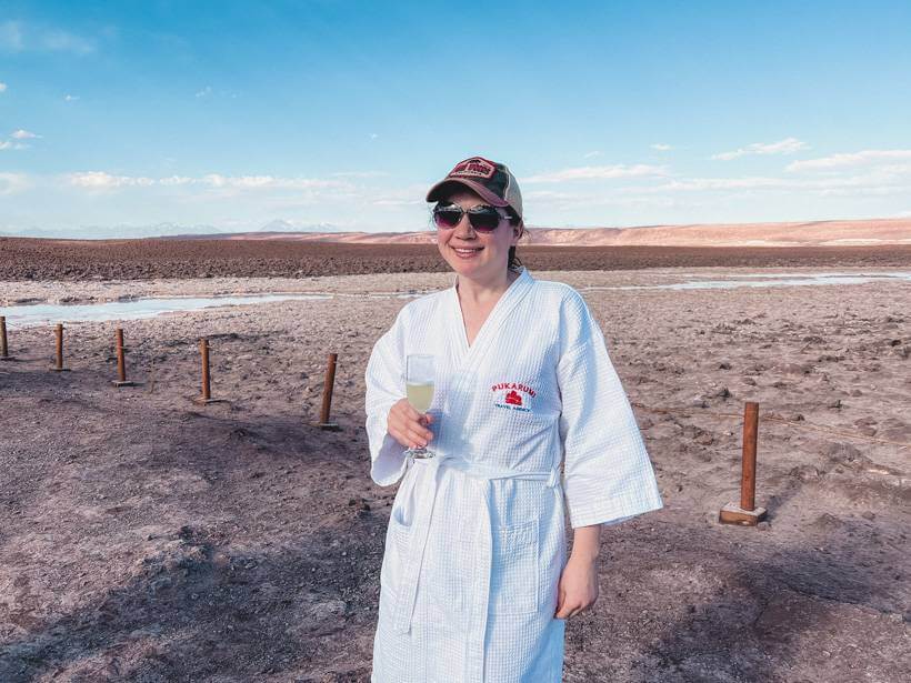 Girl holding glass of Pisco Sour in a white robe after soaking in Laguna Baltinache - Chile packing list