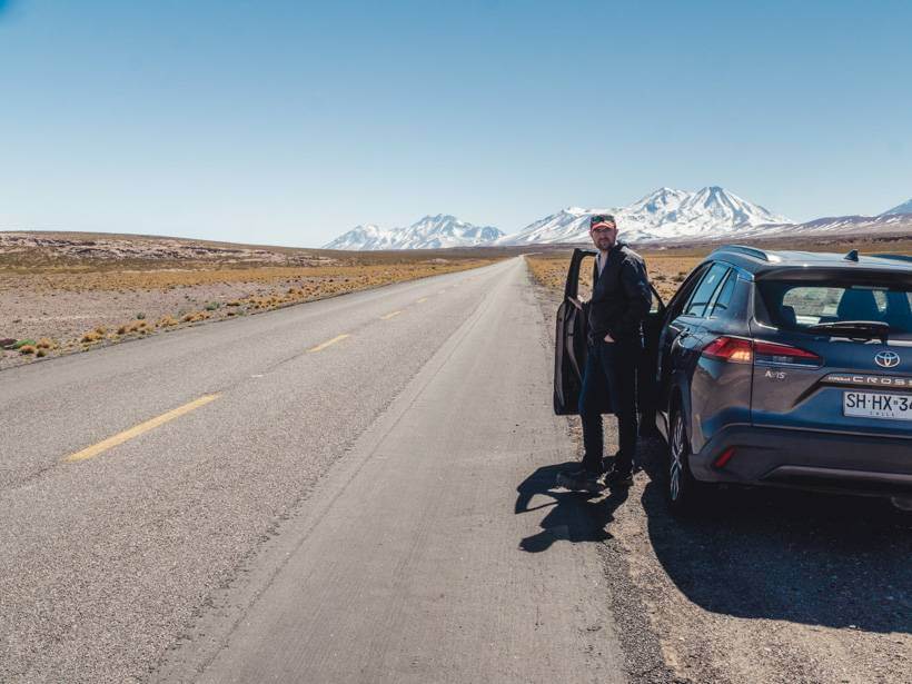 Man stands just inside car door on the side of a mountain road in the Atacama Desert with snow caps in the distance