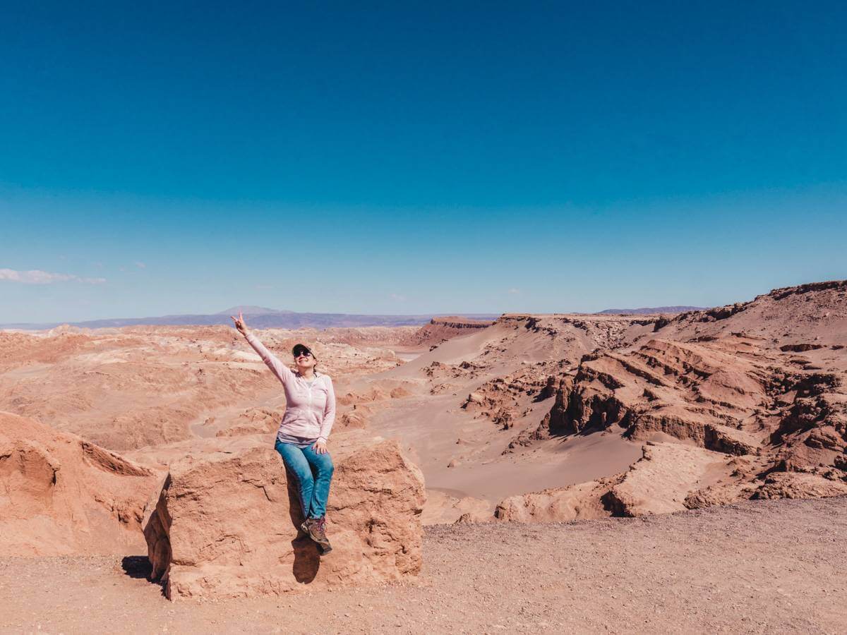 Girl in baseball cap and light pink zip-up hoodie sitting near entrance to San Pedro de Atacama during desert itinerary