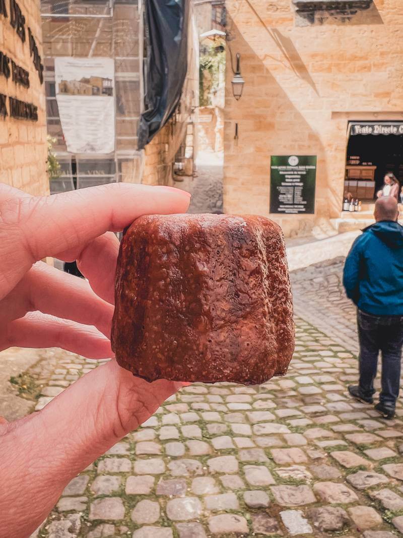 Hand holding a canelé while wandering cobblestone streets of Saint-Emilion during a day trip in Bordeaux