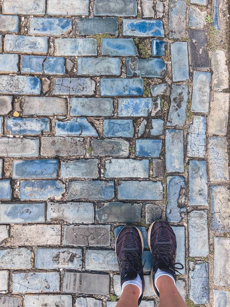 Downward view of feet wearing black sneakers standing on a street paved in blue cobbestone